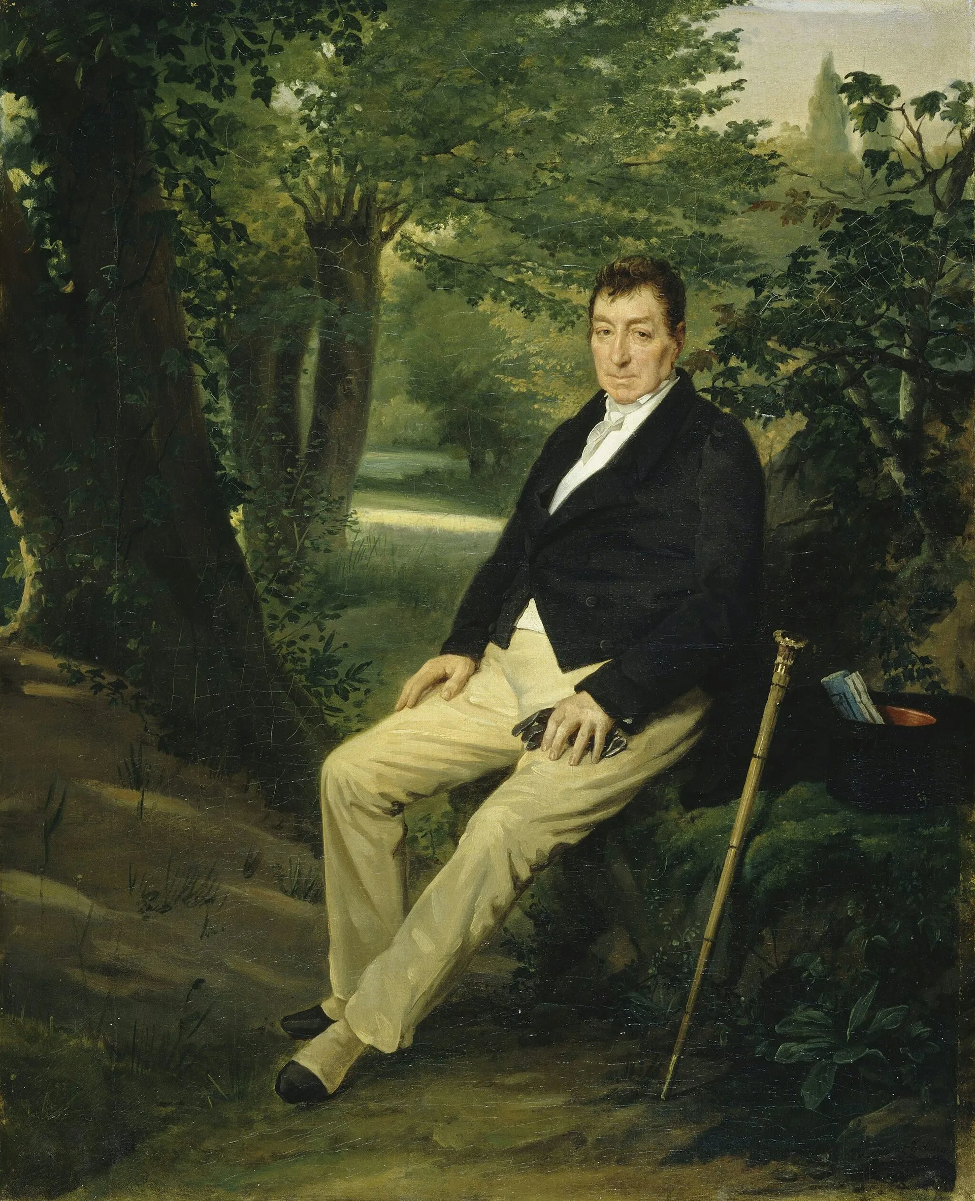 A seated, brown-haired man in a suit jacket and khaki-colored pants sits in a glen with his cane beside him